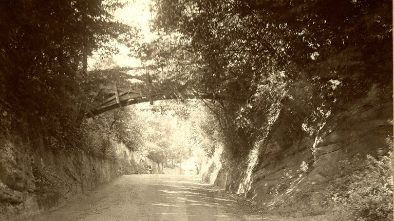 An historic photograph taken around 1900. The image looks up a road called Wightwick Bank, next to Wightwick Manor. Over the road is a wooden bridge made of straight beams, but forming a curve. Either side of the road is foliage and rock walls. In the distance on the side of the road, a few figures of people can just be seen.